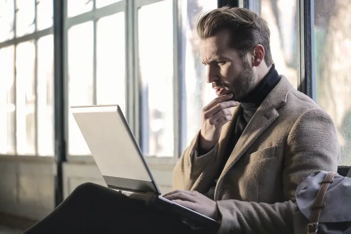 Nachdenklicher Mann sitzt mit der Hand am Kinn vor seinem Laptop am Fenster
