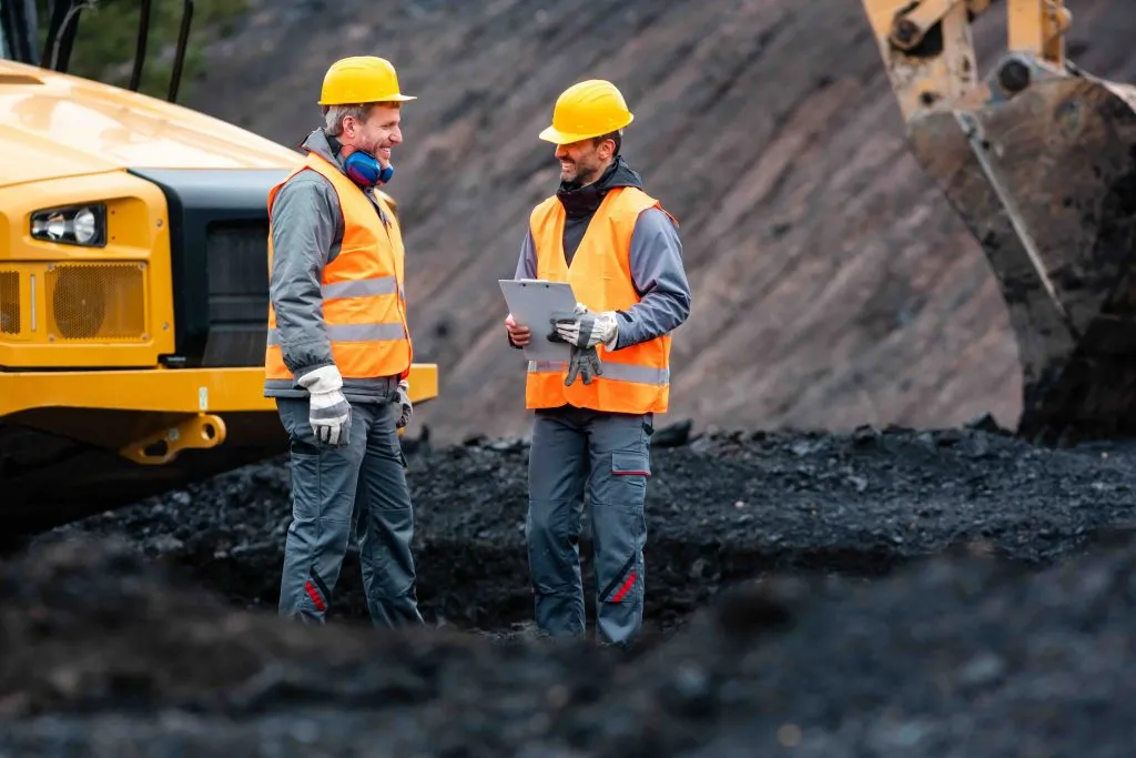 Zwei Arbeiter mit Helmen und Warnwesten besprechen sich mit Klemmbrett auf einer Baustelle vor einem Bagger