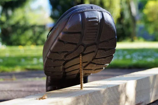 Sicherheitsschuh tritt beinahe auf eine aus einem Holzbalken ragende Schraube, typische Ursache für Arbeitsunfälle