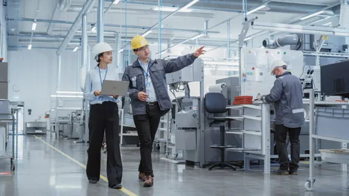 Zwei Fachkräfte mit Schutzhelmen und Laptop begehen eine Produktionshalle – Betriebssicherheit im Fokus