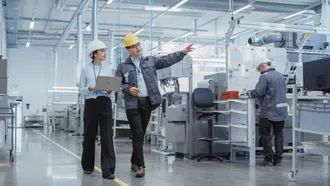 Zwei Fachkräfte mit Schutzhelmen und Laptop begehen eine Produktionshalle – Betriebssicherheit im Fokus