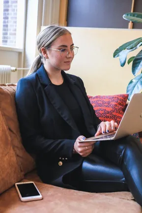 Junge Frau mit Brille sitzt auf dem Sofa und arbeitet konzentriert am Laptop