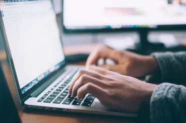 Nahaufnahme von Händen, die auf einer Laptop-Tastatur tippen, dahinter ein zweiter Monitor