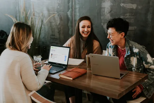 Drei lachende Mitarbeiterinnen lernen flexibel mit Laptops und einer Online-Schulungsplattform im Café