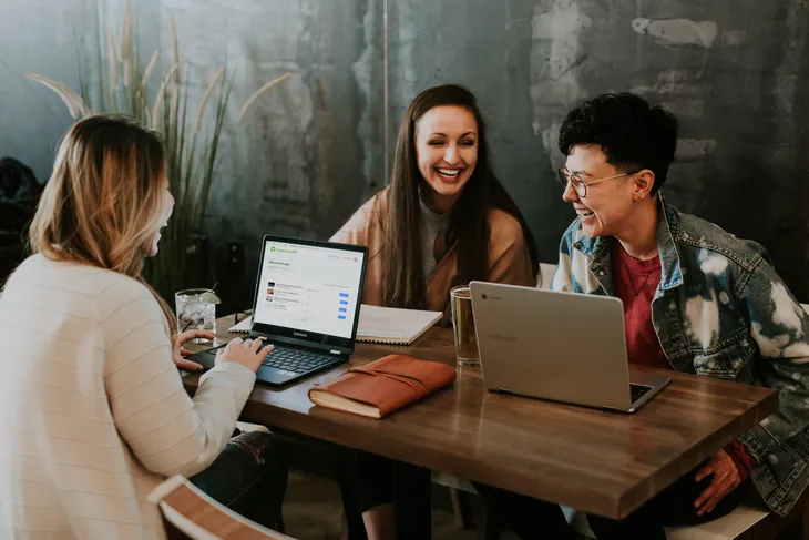 Drei lachende Mitarbeiterinnen lernen flexibel mit Laptops und einer Online-Schulungsplattform im Café