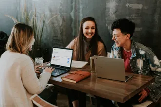 Drei lachende Mitarbeiterinnen lernen flexibel mit Laptops und einer Online-Schulungsplattform im Café