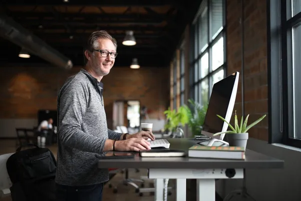 Lächelnder Mann arbeitet stehend an einem Schreibtisch mit Monitor in einem Loftbüro
