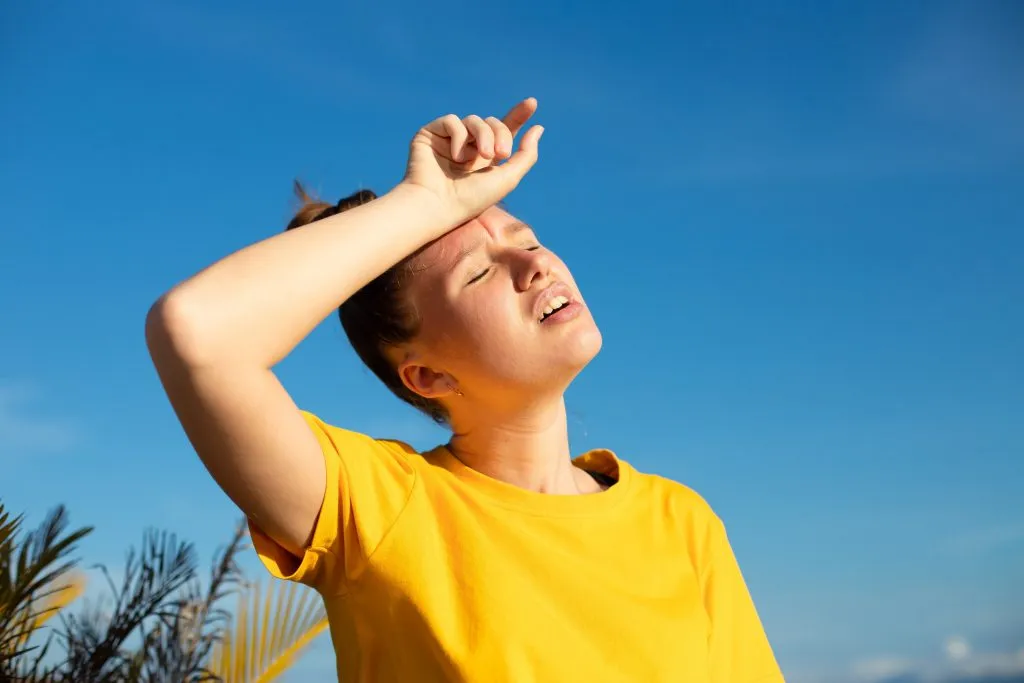Junge Frau in gelbem Shirt hält sich bei starker Sonne erschöpft den Arm an die Stirn