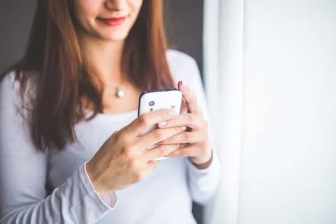 Lächelnde Frau bedient ein Smartphone mit beiden Händen am Fenster