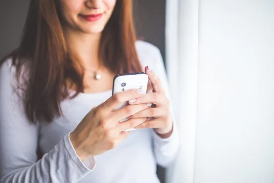 Lächelnde Frau bedient ein Smartphone mit beiden Händen am Fenster