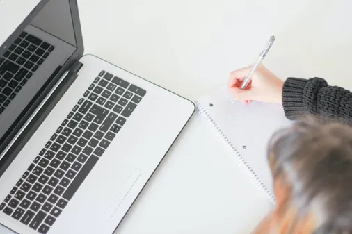 Person macht sich bei der Online Weiterbildung am Laptop handschriftliche Notizen in einen Block