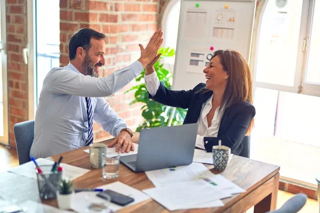 Zwei lachende Kollegen geben sich im Büro am Schreibtisch mit Laptop ein High Five