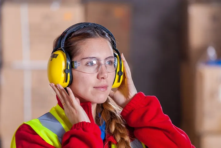 Arbeiterin mit Schutzbrille setzt gelben Gehörschutz auf, wie in der Arbeitsschutz-Unterweisung gezeigt