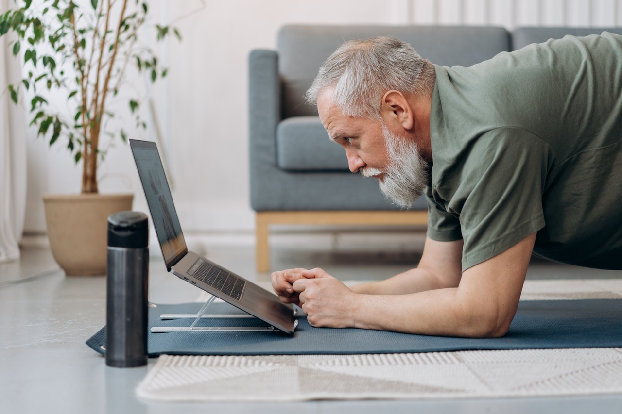 Gesundheit am Arbeitsplatz: Mann macht eine Sportübung und blickt in den Bildsdchirm eines Laptops.